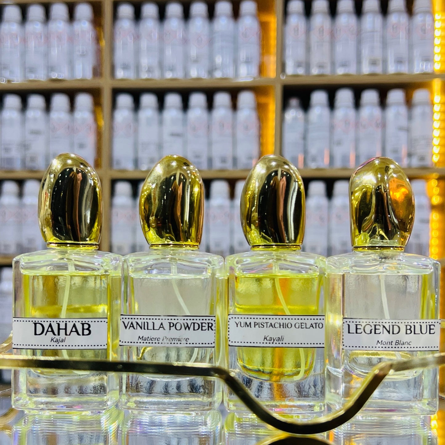 Four perfume bottles with gold caps on a reflective surface, with shelves of more bottles in the background.