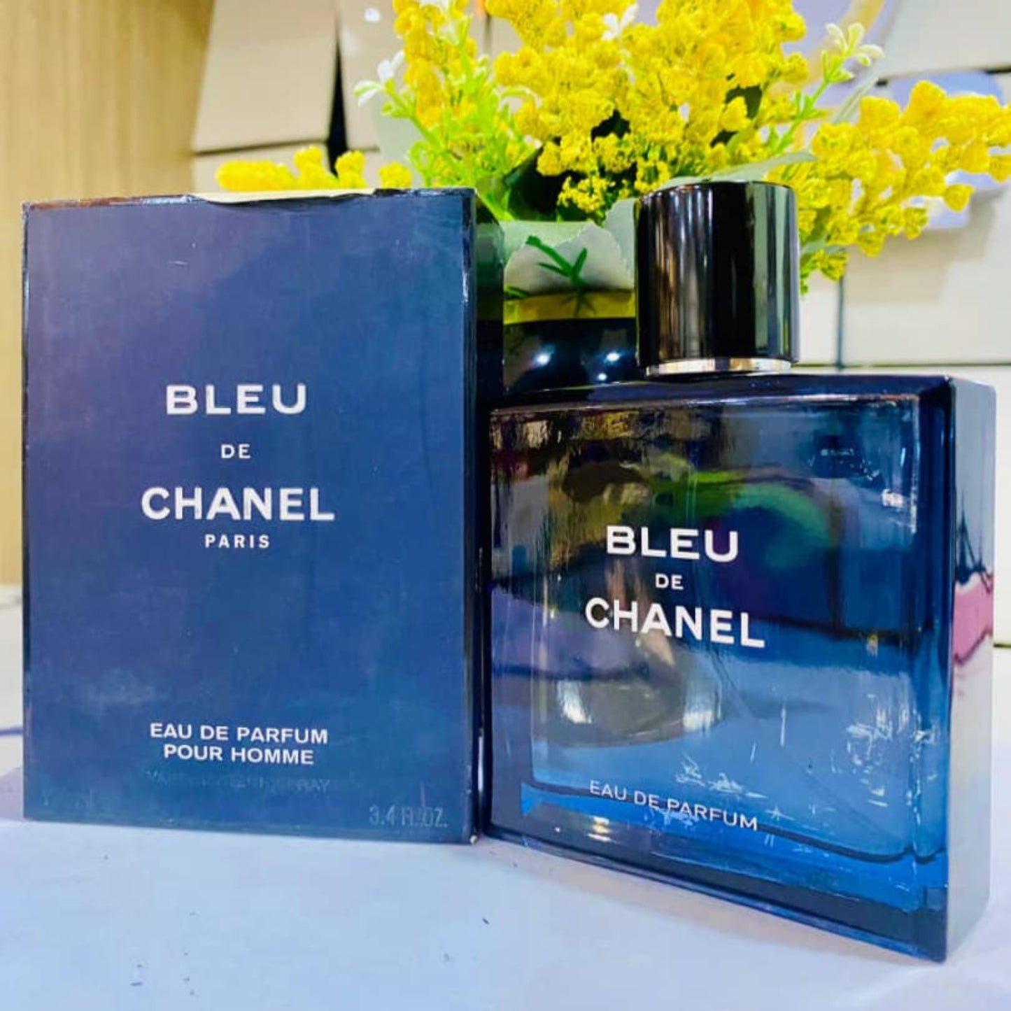 Bleu de Chanel perfume bottle and packaging on a white surface with yellow flowers in the background.
