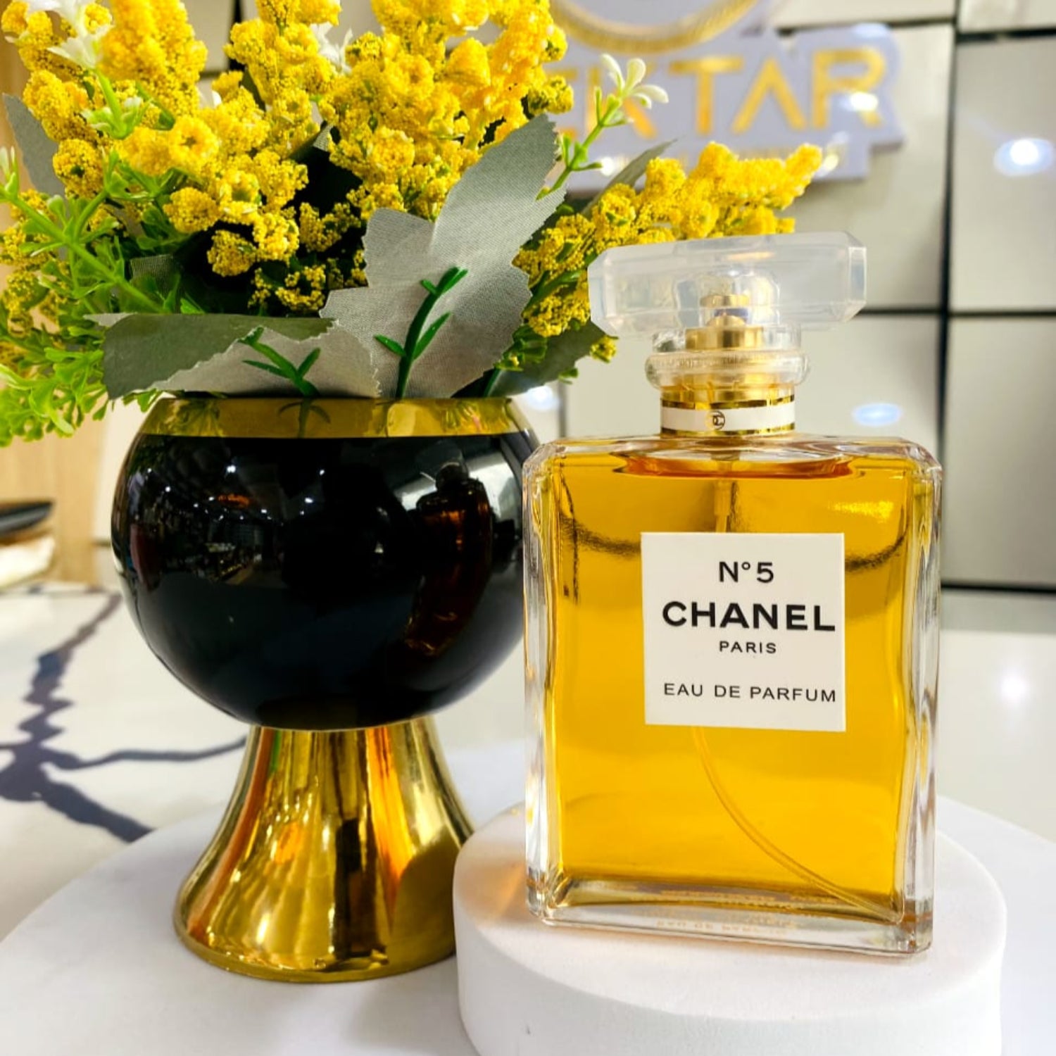 Chanel No.5 perfume bottle with yellow flowers in a black vase on a white surface.