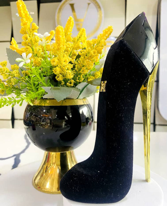 Black high-heeled shoe with gold base next to a vase with yellow flowers on a white surface.